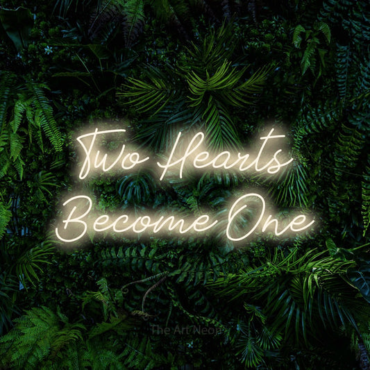 Warm white script-style LED neon sign reading "Two Hearts Become One" against a dense tropical foliage wall for wedding décor