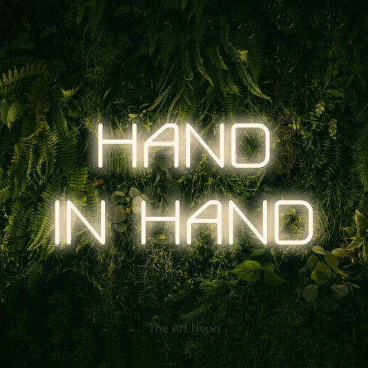 Warm white LED neon sign reading "Hand in Hand" against a moss-covered green wall for wedding décor