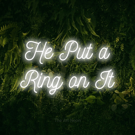Cool white script-style LED neon sign reading "He Put a Ring on It" against a moss-covered green wall for wedding décor