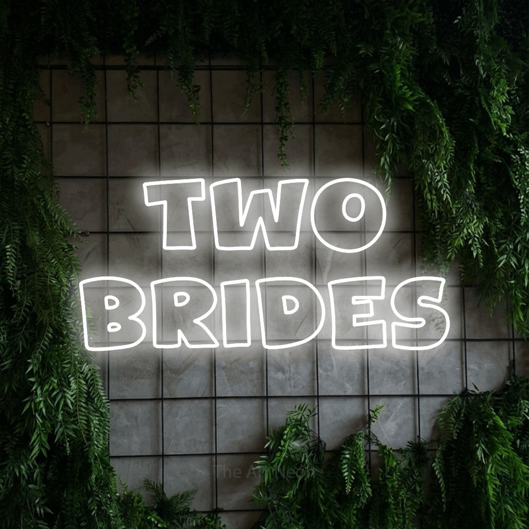 Cool white outline-style LED neon sign reading "Two Brides" against a green grid wall with foliage for wedding décor