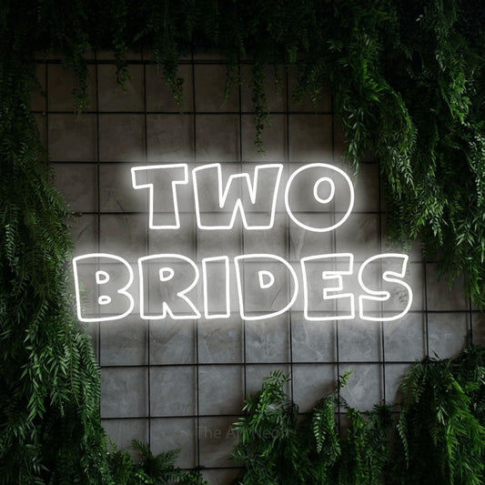 Cool white outline-style LED neon sign reading "Two Brides" against a green grid wall with foliage for wedding décor