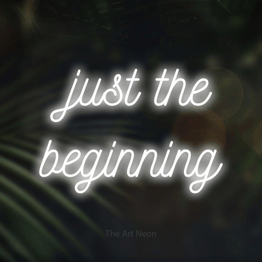 Cool white script-style LED neon sign reading "Just the Beginning" against dark blurred palm leaves for wedding décor