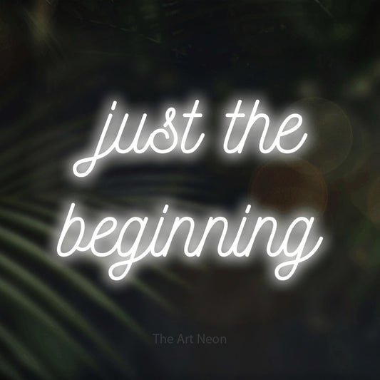 Cool white script-style LED neon sign reading "Just the Beginning" against dark blurred palm leaves for wedding décor