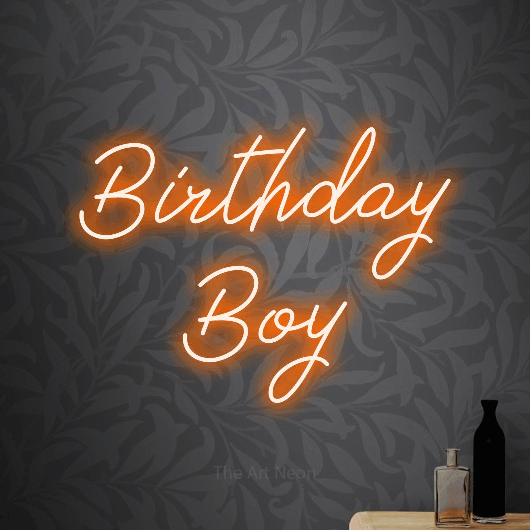 Orange script-style LED neon sign reading "Birthday Boy" against light grey floral wallpaper for party décor