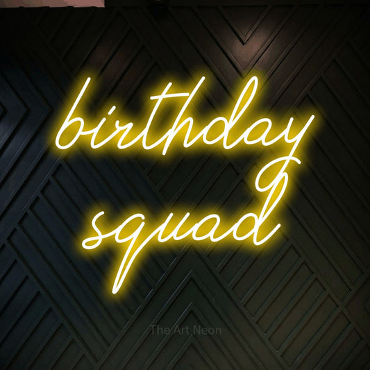 Yellow script-style LED neon sign reading "Birthday Squad" against a dark geometric wood wall for party décor
