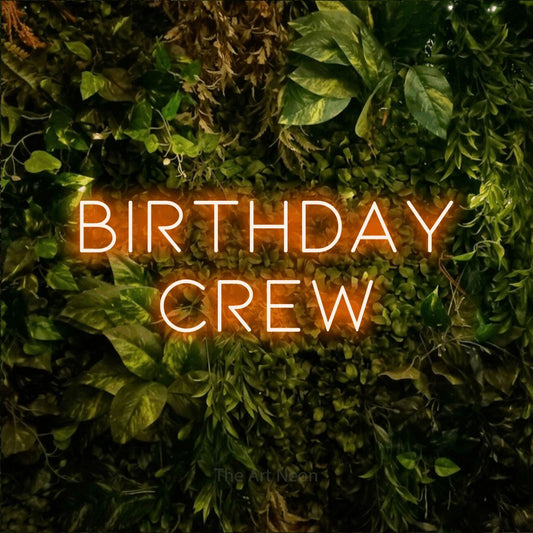 Orange LED neon sign reading "Birthday Crew" against a textured moss and foliage wall for party décor