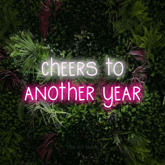 Hot pink and cool white LED neon sign reading "Cheers to Another Year" against a lush garden wall with pink flowers