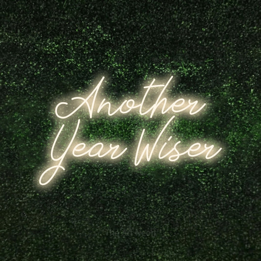 Warm white script-style LED neon sign reading "Another Year Wiser" against a textured green moss wall for party décor