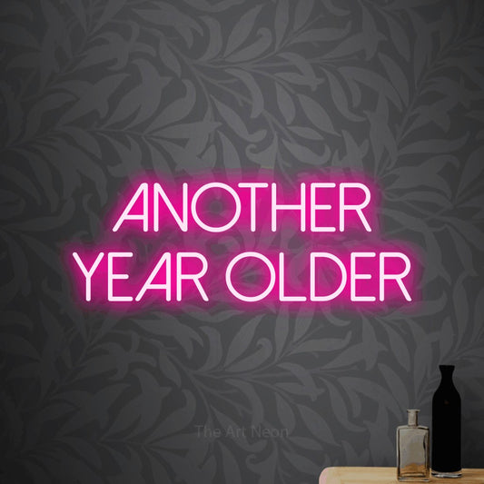 Hot pink LED neon sign reading "Another Year Older" against light grey floral wallpaper for party décor