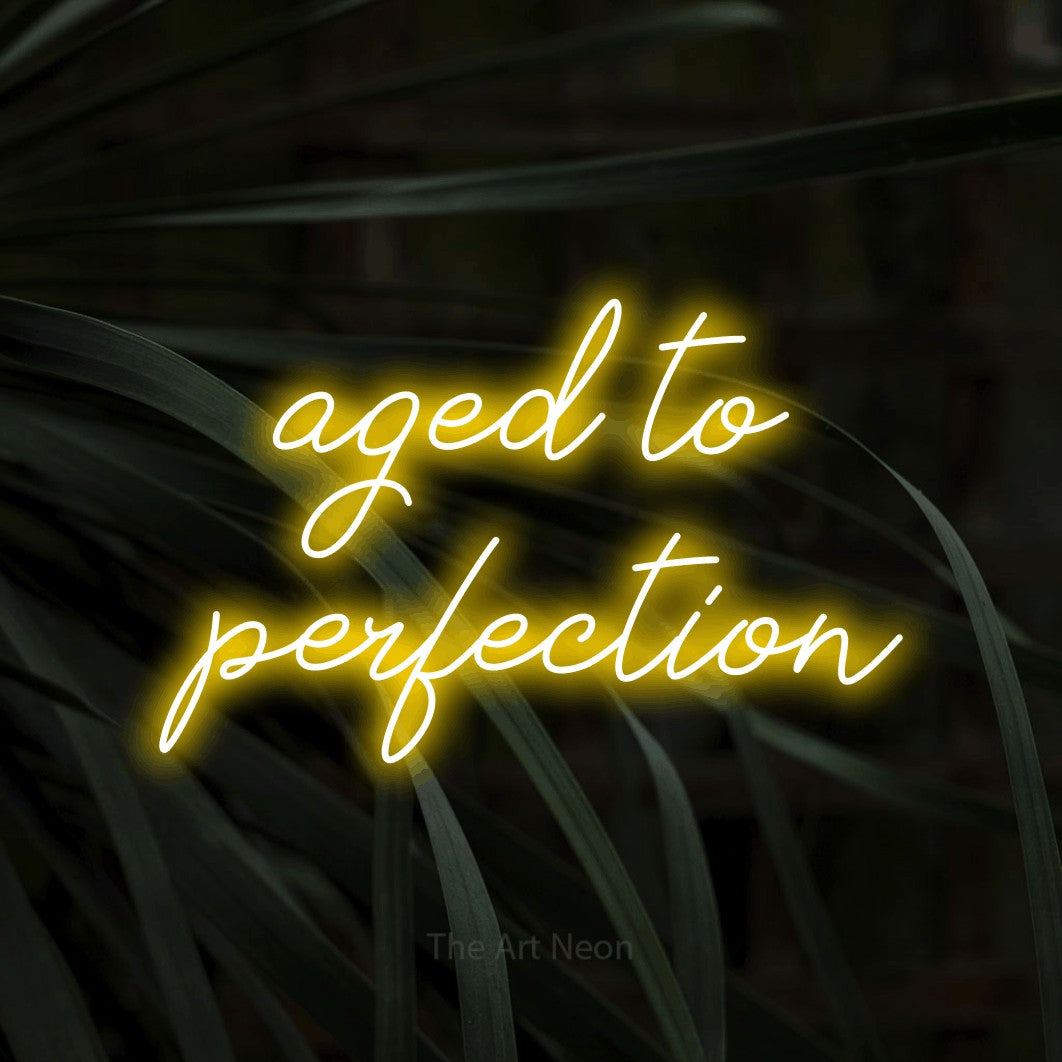 Enseigne néon à DEL à lumière jaune, de style manuscrit, affichant « Aged to Perfection » sur des feuilles tropicales foncées, pour décorer une fête