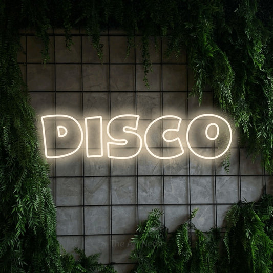 Warm white outline-style LED neon sign reading "Disco" against a green grid wall with foliage for party décor