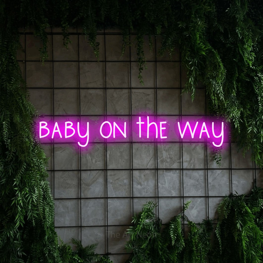 Enseigne néon à DEL à lumière rose, affichant « Baby on the Way » sur un mur quadrillé vert avec du feuillage