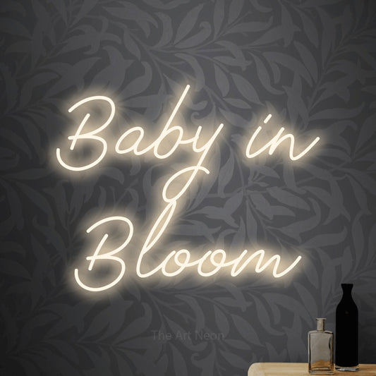 Warm white script-style LED neon sign reading "Baby in Bloom" against light grey floral wallpaper