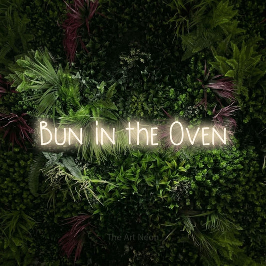 Warm white LED neon sign reading "Bun in the Oven" against a lush garden wall with pink flowers