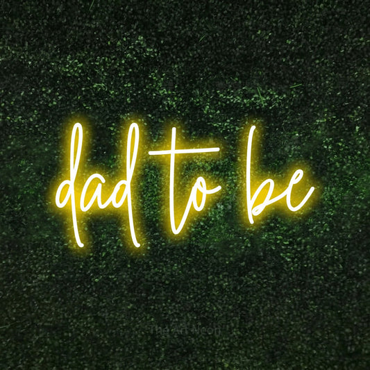 Enseigne néon à DEL à lumière jaune, de style manuscrit, affichant « Dad to Be » sur un mur texturé de mousse verte