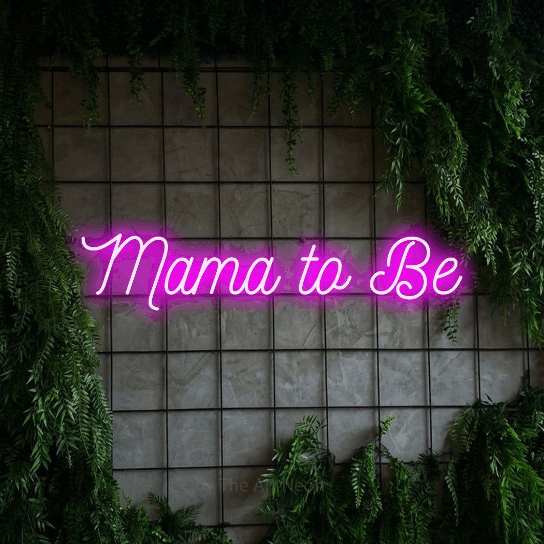 Pink script-style LED neon sign reading "Mama to Be" against a green grid wall with foliage