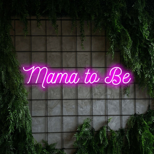 Enseigne néon à DEL à lumière rose, de style manuscrit, affichant « Mama to Be » sur un mur quadrillé vert avec du feuillage