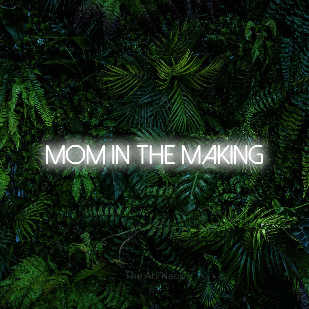 Cool white LED neon sign reading "Mom in the Making" against a dense tropical foliage wall