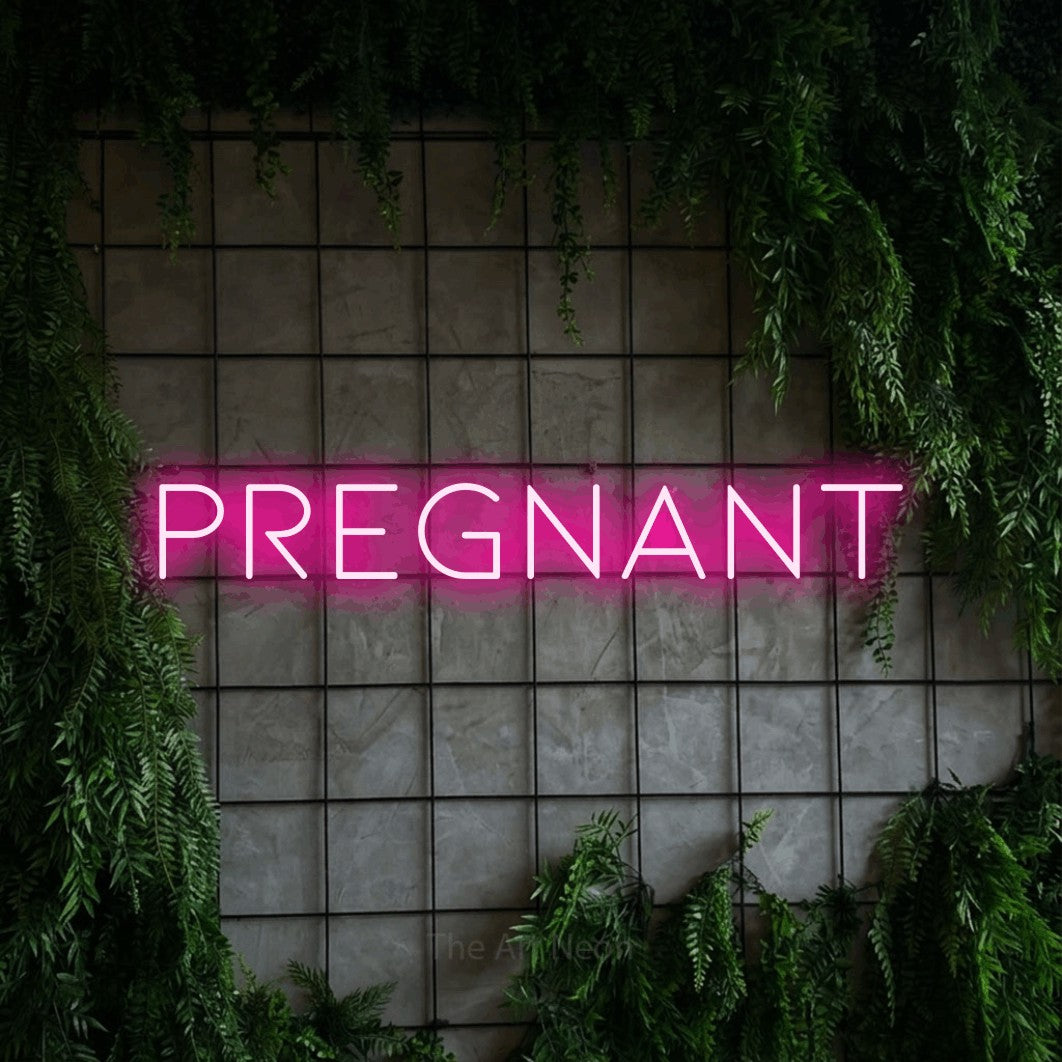 Hot pink LED neon sign reading "Pregnant" against a green grid wall with foliage