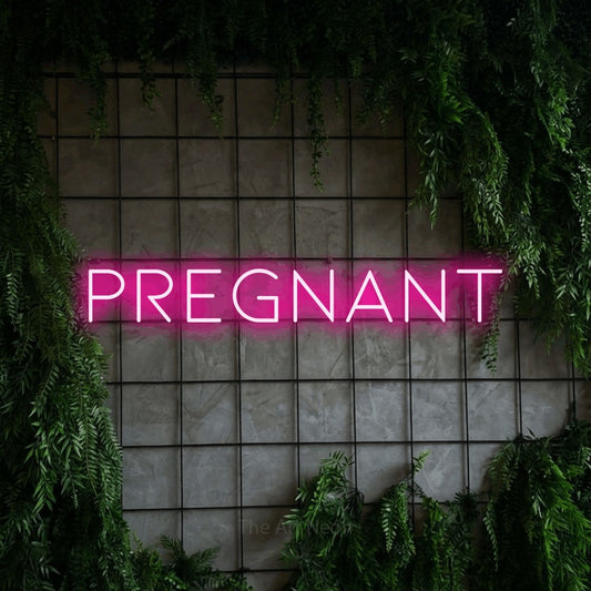 Enseigne néon à DEL à lumière rose vif, affichant « Pregnant » sur un mur quadrillé vert avec du feuillage