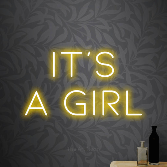 Yellow LED neon sign reading "It's a Girl" against light grey floral wallpaper