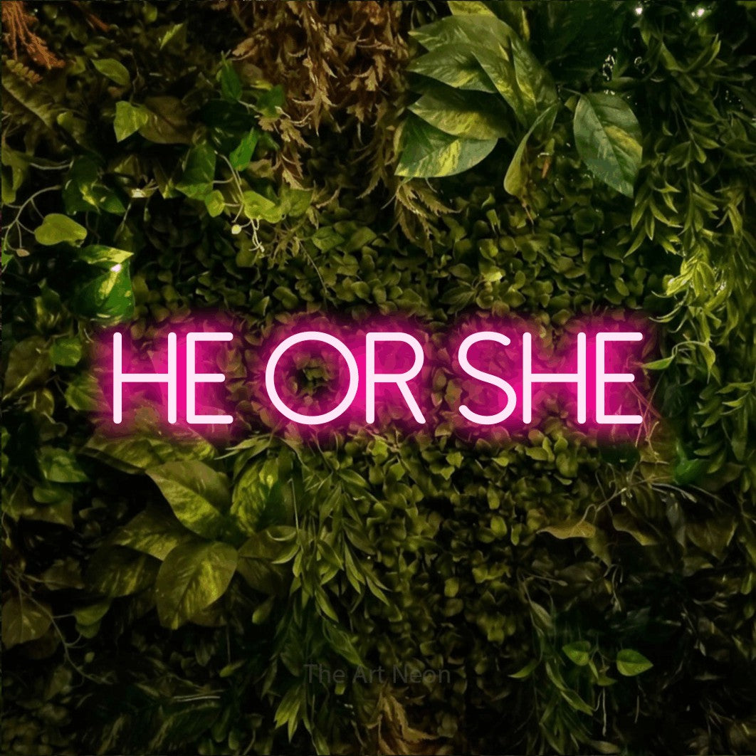 Hot pink LED neon sign reading "He or She" against a textured moss and foliage wall