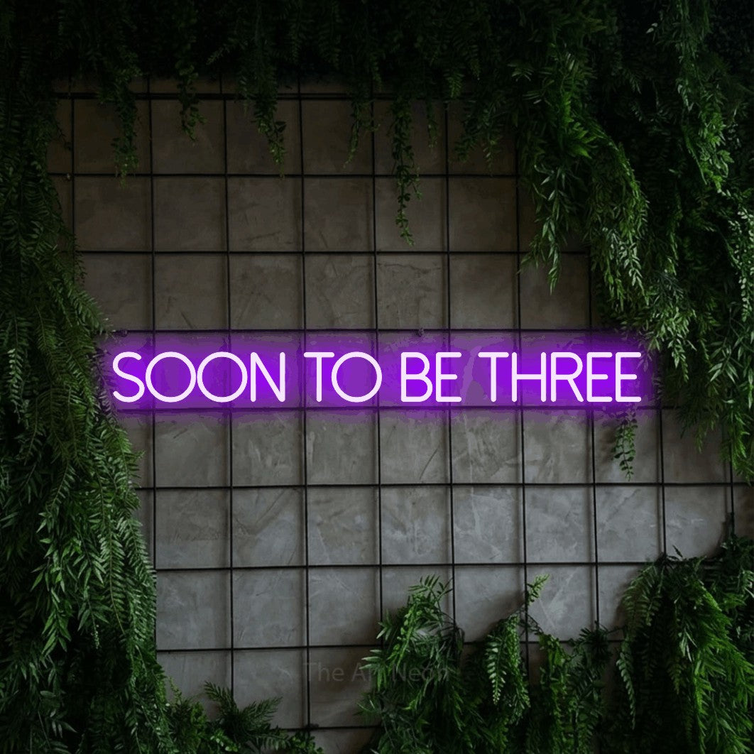 Enseigne néon à DEL à lumière violette, affichant « Soon to Be Three » sur un mur quadrillé vert avec du feuillage