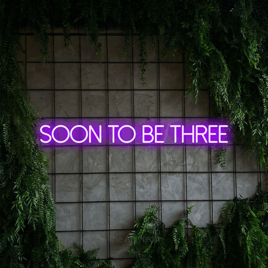 Purple LED neon sign reading "Soon to Be Three" against a green grid wall with foliage