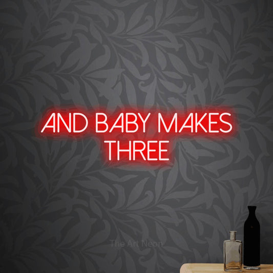 Red LED neon sign reading "And Baby Makes Three" against light grey floral wallpaper