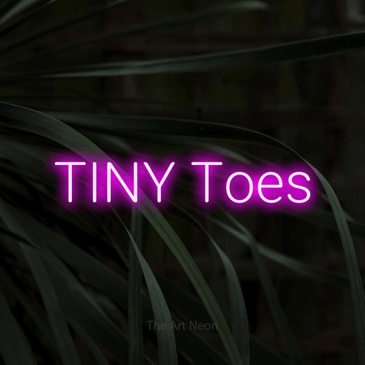 Pink LED neon sign reading "Tiny Toes" against dark tropical leaves