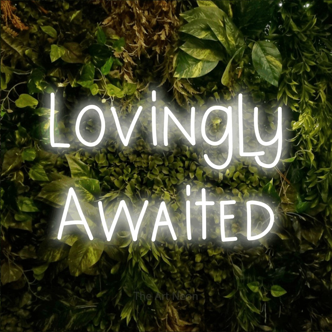 Cool white LED neon sign reading "Lovingly Awaited" against a textured moss and foliage wall