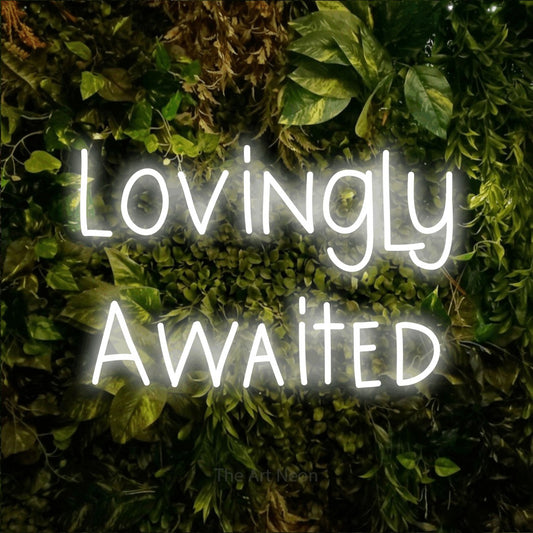 Cool white LED neon sign reading "Lovingly Awaited" against a textured moss and foliage wall