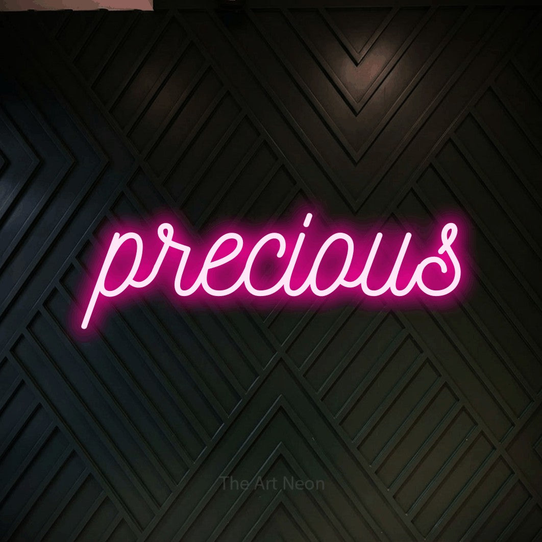 Hot pink script-style LED neon sign reading "Precious" against a dark geometric wood wall
