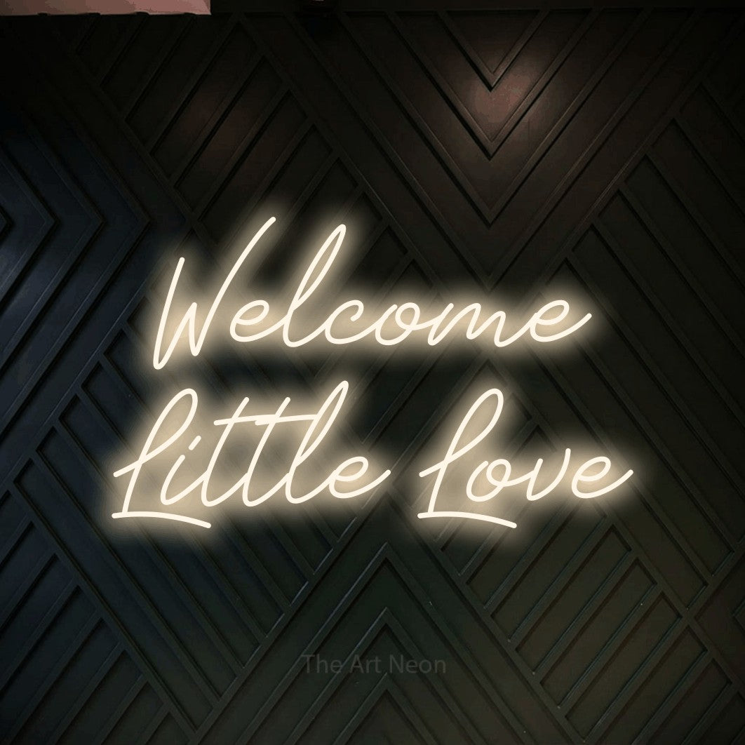 Warm white script-style LED neon sign reading "Welcome Little Love" against a dark geometric wood wall