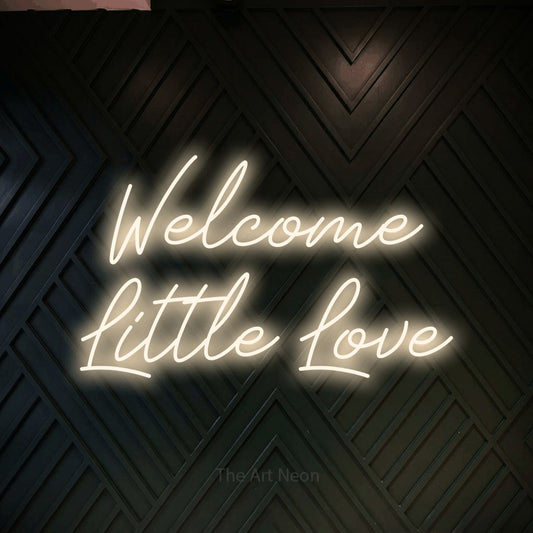 Warm white script-style LED neon sign reading "Welcome Little Love" against a dark geometric wood wall