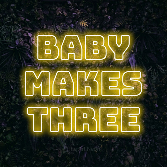 Yellow outline-style LED neon sign reading "Baby Makes Three" against a purple floral wall