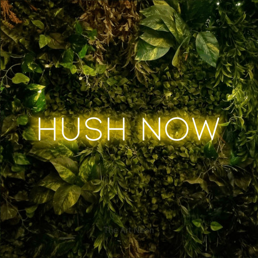 Yellow LED neon sign reading "Hush Now" against a textured moss and foliage wall