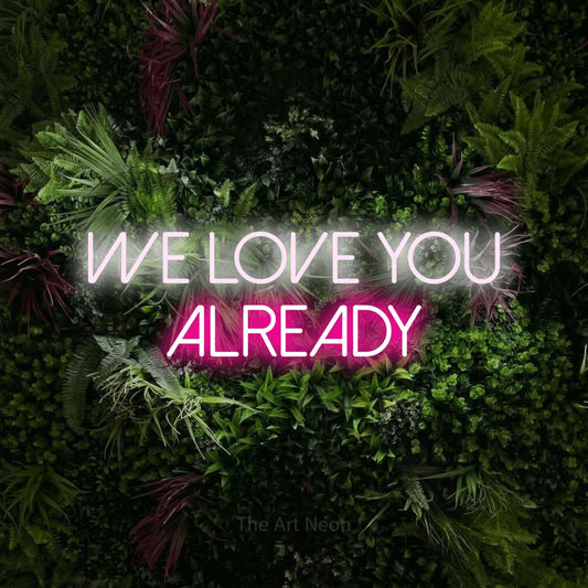 Hot pink and cool white LED neon sign reading "We Love You Already" against a lush garden wall with pink flowers