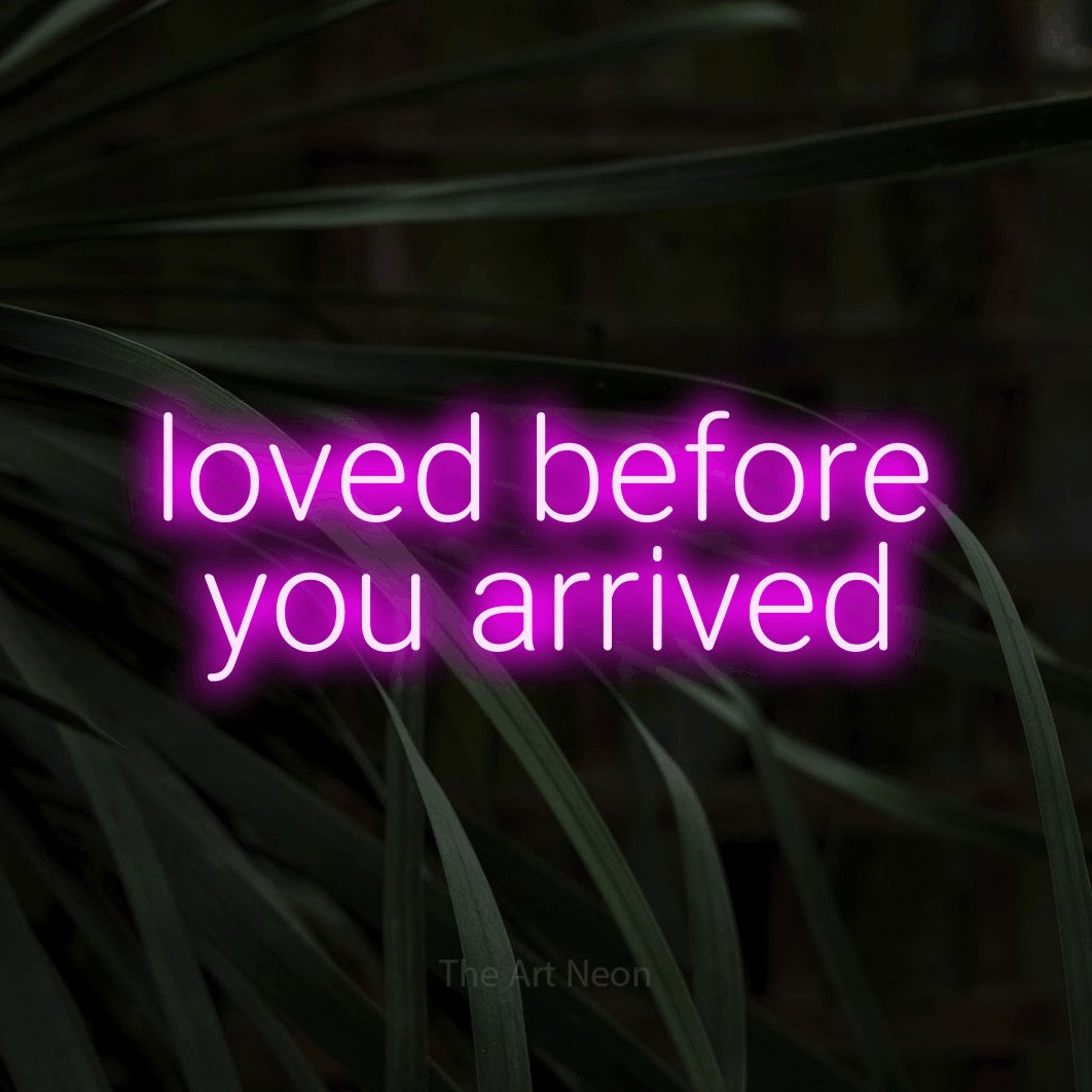 Pink LED neon sign reading "Loved Before You Arrived" against dark tropical leaves