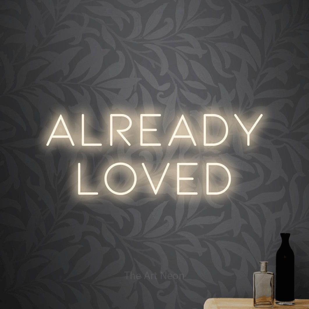 Warm white LED neon sign reading "Already Loved" against light grey floral wallpaper