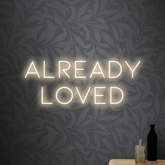 Warm white LED neon sign reading "Already Loved" against light grey floral wallpaper