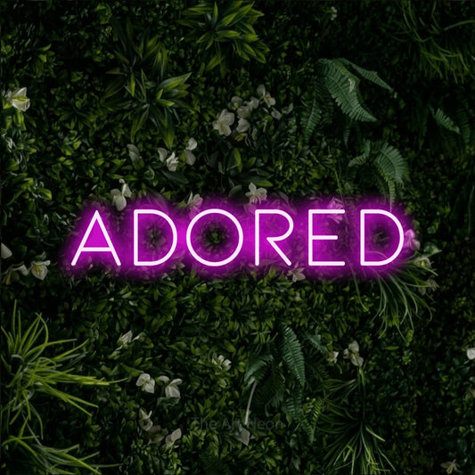 Pink LED neon sign reading "Adored" against a leafy green wall