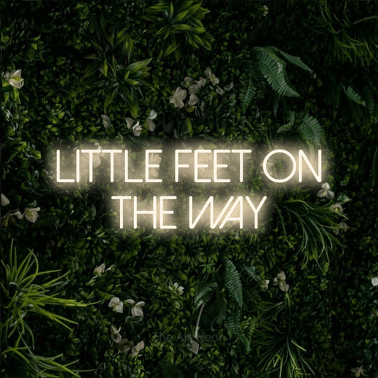 Warm white LED neon sign reading "Little Feet on the Way" against a leafy green wall