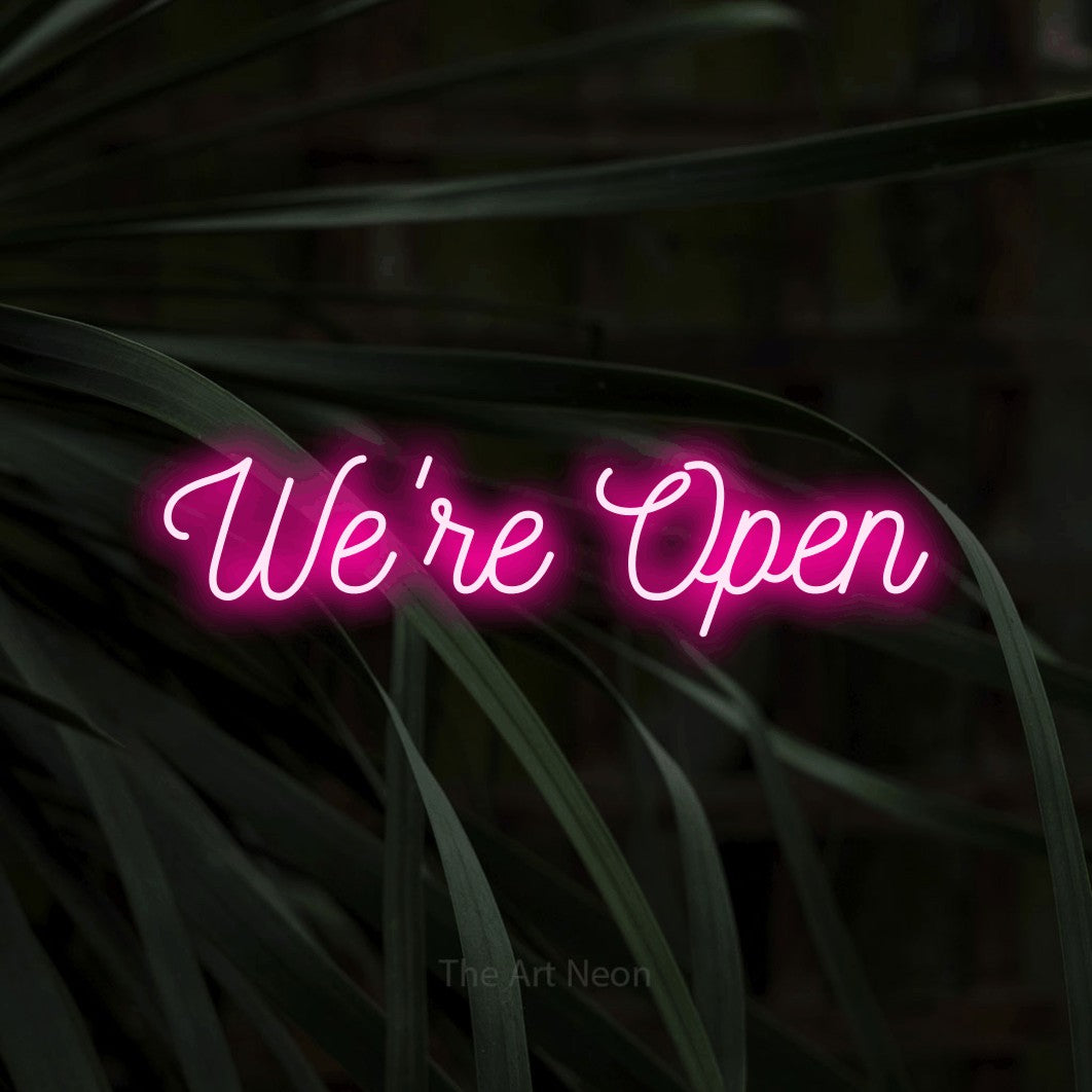 Hot pink script-style LED neon sign reading "We're Open" against dark tropical leaves for restaurant décor