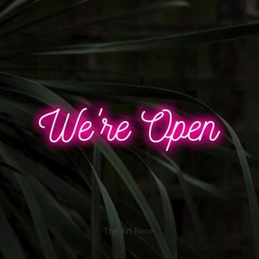 Hot pink script-style LED neon sign reading "We're Open" against dark tropical leaves for restaurant décor