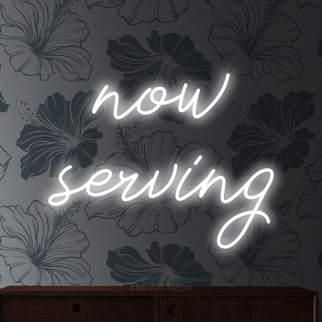 Cool white script-style LED neon sign reading "Now Serving" against grey floral wallpaper for restaurant décor
