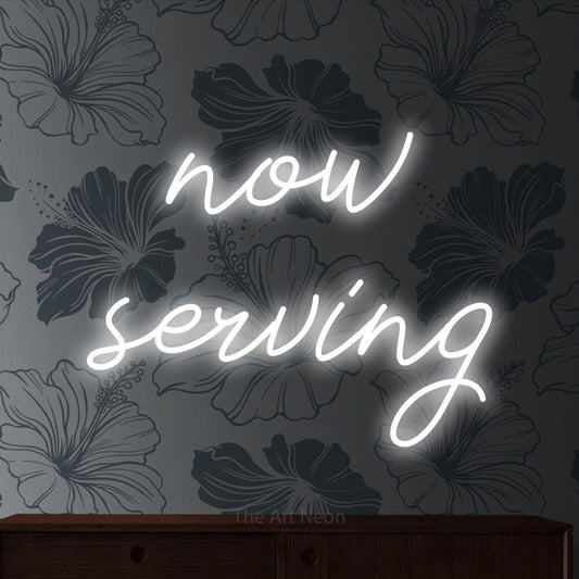 Cool white script-style LED neon sign reading "Now Serving" against grey floral wallpaper for restaurant décor
