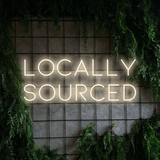 Warm white LED neon sign reading "Locally Sourced" against a green grid wall with foliage for restaurant décor
