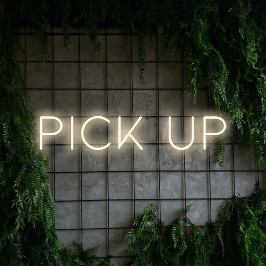 Warm white LED neon sign reading "Pick Up" against a green grid wall with foliage for restaurant décor
