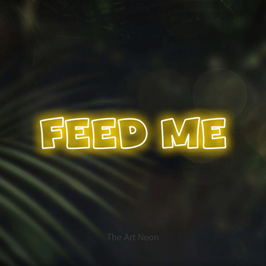 Yellow outline-style LED neon sign reading "Feed Me" against dark blurred palm leaves for restaurant décor